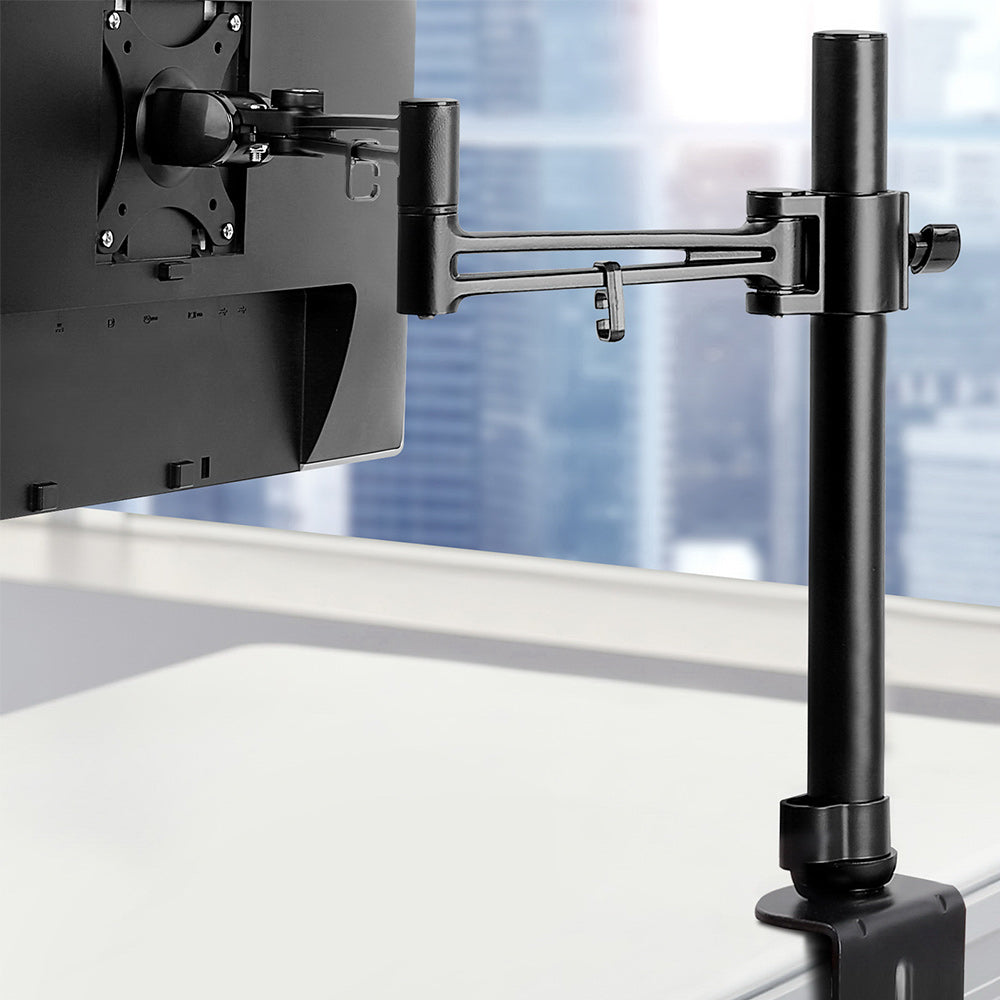 Black monitor stand with a monitor attached, set against a blurred office window background.