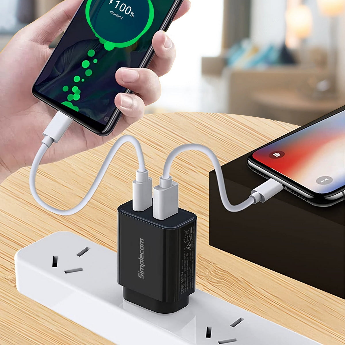 Person holding a smartphone with a charging cable connected to a power adapter on a wooden surface.